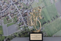 Zu sehen ist der Wanderpokal in Form eines Radfahrers.  Im Hintergrund ist ein Foto von oben von Straßen und Feldern zu sehen.