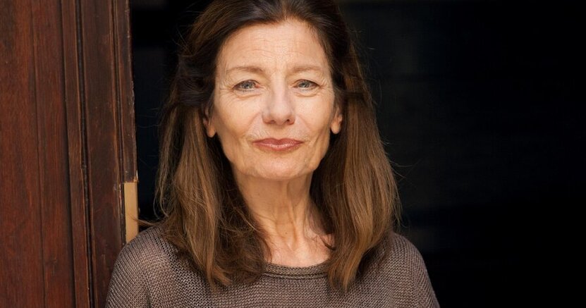 Ursula Krechel hat ein braunes Oberteil mit kurzen Ärmeln an und lehnt sich an einen dunkelbraunen Türrahmen.