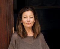 Ursula Krechel hat ein braunes Oberteil mit kurzen Ärmeln an und lehnt sich an einen dunkelbraunen Türrahmen.