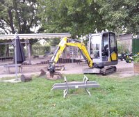 Zu sehen ist ein gelber Bagger auf Sand. Er steht neben einem Rasen.