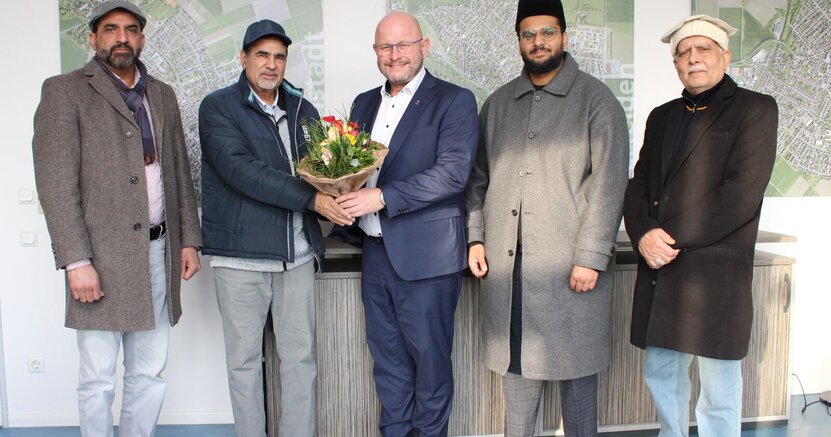 Atta-ul Ghafoor, Abdul Shakoor, Bürgermeister Marcus Kretschmann, Imam Awayes Ahmad Malik und Mumtaz Ahmad (v.li.)stehen im Arbeitszimmer des Bürgermeisters vor Wandbildern, die Luftaufnahmen der Stadtteile zeigen. Der Bürgermeister hat einen Blumenstrauß in der Hand.