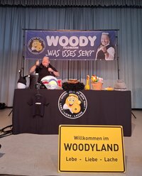 Woody Feldmann steht hinter ihrem Bühnentisch und reckt den Daumen nach oben. Vor ist ein großes gelbes Ortsschild mit der Aufschrift "Woodyland" zu sehen. Über ihr befindet sich ein großes Banner mit ihrem Namen.