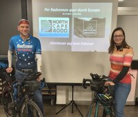 Tom und Stevie Thissen stehen mit ihren Fahrrädern im Saal des alten Rathauses Crumstadt. Zwischen ihnen befindet sich eine Leinwand. Drauf ist die Ankündigung ihres Vortrags zu lesen.