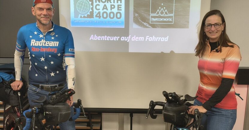 Tom und Stevie Thissen stehen mit ihren Fahrrädern im Saal des alten Rathauses Crumstadt. Zwischen ihnen befindet sich eine Leinwand. Drauf ist die Ankündigung ihres Vortrags zu lesen.
