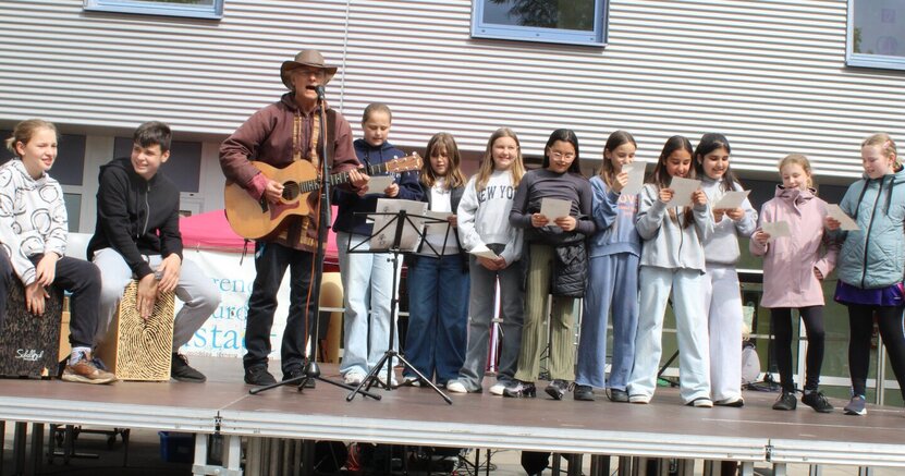 Uwe Schatter sang mit Schülerinnen und Schülern der MNS das neue Kinderfest-Lied Uwe Schatter steht mit Hut und seiner Gitarre auf der Bühne des Kinderfestes. Vor sich hat er ein Mikorofon. Schülerinnen und Schüler stehen in einer Reihe hinter ihm und halten Zettel. Sie singen alle gemeinsam das neue Kinderfest-Lied.