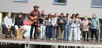 Uwe Schatter sang mit Schülerinnen und Schülern der MNS das neue Kinderfest-Lied Uwe Schatter steht mit Hut und seiner Gitarre auf der Bühne des Kinderfestes. Vor sich hat er ein Mikorofon. Schülerinnen und Schüler stehen in einer Reihe hinter ihm und halten Zettel. Sie singen alle gemeinsam das neue Kinderfest-Lied.