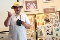 Forscher Dr. Cornelius Gitterbaum in seinem Element. Forscher Dr. Cornelius Gitterbaum hat einen Tropenhelm auf. Um seinen Hals hängt ein Fernglas. Er reckt einen Daumen hoch. Neben ihm sieht man eine Tafel mit Abbildungen von Schneckengehäusen, Vögeln, Keramikgefäßen und anderem.