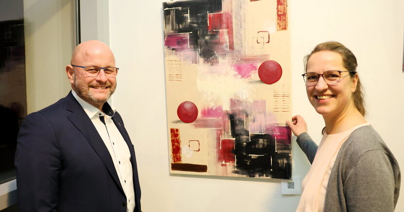 Bürgermeister Marcus Kretschmann und Künstlerin Aida Hiseni stehen links und rechts vor einem Werk der Künstlerin. Es ist ein abstraktes Gemälde mit bunten Kugeln und rechteckigen Formen.