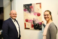 Bürgermeister Marcus Kretschmann und Künstlerin Aida Hiseni stehen links und rechts vor einem Werk der Künstlerin. Es ist ein abstraktes Gemälde mit bunten Kugeln und rechteckigen Formen.