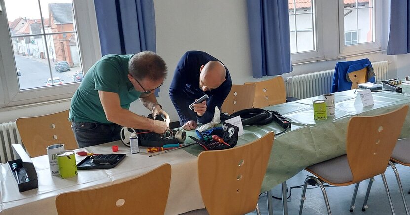 Zwei Männer beugen sich an einem Tisch über ein elektrisches Gerät. Der eine Mann leuchtet mit einer Taschenlampe, während der andere versucht, das Gerät zu reparieren. 