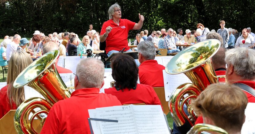 Im Vordergrund sind Musiker des Musikvereins in roten T-Shirts zu sehen. Sie sitzen mit dem Rücken zum Betrachter. Vor ihnen steht Dirigent Thomas Grimm. Im Hintergrund sind Zuschauer auf Bierbänken im Volkspark Goddelau zu sehen. Hinter ihnen stehen Bäume.
