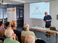 Rudolf Bersch erklärt die Energiewende. Rudolf Bersch steht an einem Tisch im Sitzungssaal des Rathauses. Hinter ihm ist an der Wand eine Grafik aus seinem Vortrag zu sehen. Vor ihm sitzen Zuschauer und hören ihm zu.