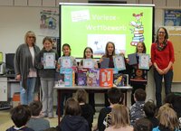 Schulleiterin Susanne Möller mit Marie, Betty, Julia, Luise, der Erstplatzierten Theresa und Büchereileiterin Anja Stark (v.li.). Schulleiterin Susanne Möller steht mit Marie, Betty, Julia, Luise, der Erstplatzierten Theresa und Büchereileiterin Anja Stark (v.li.)vor den auf dem Boden sitzenden Viertklässlern. Die Vorlese-Kinder halten ihre Urkunden hoch. Im Hintergrund ist auf einer Leinwand zu lesen: "Vorlesewettbewerb 2026"