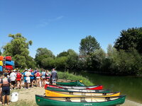 Links von einem Fluss liegen auf dem Ufer mehrere Kanus. Jugendliche in Schwimmwesten stehen daneben, mit dem Rücken zum Betrachter.