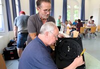 Im Vordergrund schrauben zwei Männer an einem Gerät. Im Hintergrund sind weitere Menschen an Tischen zu sehen.