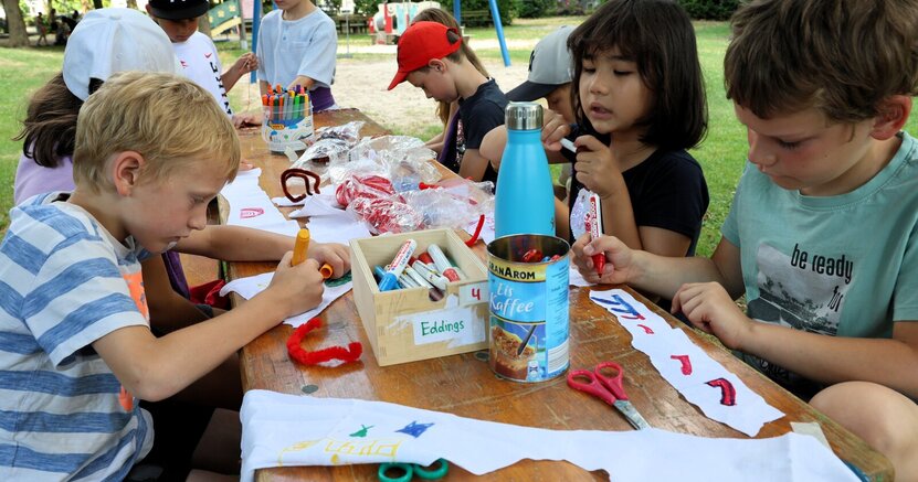 Ferienspielkinder bemalen im Volkspark Goddelau Stirnbänder. Kinder sitzen im Volkspark an einem langen Tisch. Sie bemalen mit bunten Stiften weiße Tuchstreifen.