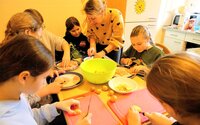 Kinder und Jugendliche sitzen um einen Tisch herum und schneiden Zutaten für das gemeinsame Essen. Jugendpflegerin Julia Spitzer hilft.