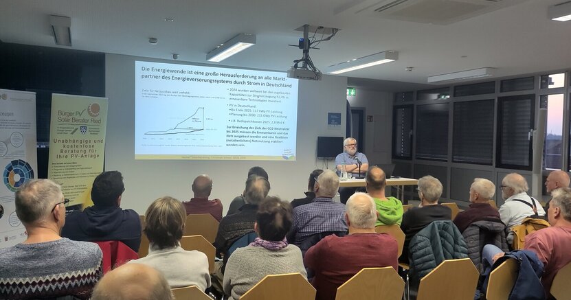 Referent Christoph Schaaf sitzt im Sitzungssaal des Riedstädter Rathauses vor einem großen Publikum an einem Tisch. Neben ihm ist auf einer Leinwand an der Wand eine Seite seiner Präsentation mit einer Grafik zu sehen.