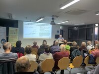 Referent Christoph Schaaf erklärt dynamische Stromtarife und variable Netz-entgelte. Referent Christoph Schaaf sitzt im Sitzungssaal des Riedstädter Rathauses vor einem großen Publikum an einem Tisch. Neben ihm ist auf einer Leinwand an der Wand eine Seite seiner Präsentation mit einer Grafik zu sehen.