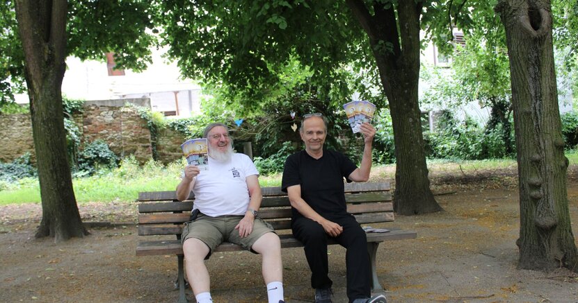 Werner Schmidt und Christian Suhr (v.li.) präsentieren „Unter den Linden“ das Programm des Büchnerland-Festivals Werner Schmidt und Christian Suhr sitzen auf einer Bank unter mächtigen Linden. Sie halten jeweils mehrere Flyer mit dem Programm des Büchner-Landfestivals hoch.