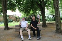 Werner Schmidt und Christian Suhr sitzen auf einer Bank unter mächtigen Linden. Sie halten jeweils mehrere Flyer mit dem Programm des Büchner-Landfestivals hoch.