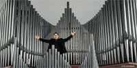 Bernhardt Brand-Hofmeister steht mit ausgebreiteten Armen vor einer Orgel
