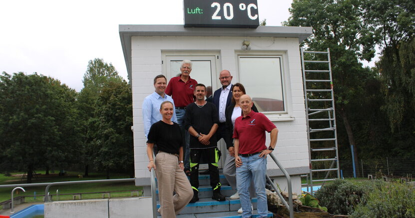 Inna Wedel, Alexander Schaper, Werner Amend, Sascha Schmidt, Bürgermeister Marcus Kretschmann, Dorota Bayram und Holger Michels stehen im Freibad Goddelau auf einer Treppe mit blauen Stufen. Die Treppe führt zu einem weiß gestrichenen Turm. Auf dem Turm befindet sich die neue Uhr. Sie zeigt gerade an "Luft: 20 Grad"