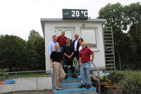 Inna Wedel, Alexander Schaper, Werner Amend, Sascha Schmidt, Bürgermeister Marcus Kretschmann, Dorota Bayram und Holger Michels (v.li.) unter der neuen Schwimmbaduhr. Inna Wedel, Alexander Schaper, Werner Amend, Sascha Schmidt, Bürgermeister Marcus Kretschmann, Dorota Bayram und Holger Michels stehen im Freibad Goddelau auf einer Treppe mit blauen Stufen. Die Treppe führt zu einem weiß gestrichenen Turm. Auf dem Turm befindet sich die neue Uhr. Sie zeigt gerade an "Luft: 20 Grad"