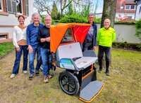 Nicole Novak, Manfred Rachut, Horst Geißler, Bernd Rupp und Alois Ittner (v.li.) freuen sich auf die neue Rikscha-Saison Nicole Novak, Manfred Rachut, Horst Geißler, Bernd Rupp und Alois Ittner stehen draußen neben dem Gebäude der Stiftung Soziale Gemeinschaft Riedstadt. Vor ihnen steht die Fahrrad-Rikscha mit orangem Verdeck und grauen Sitzpolstern sowie grauer Fußbank.