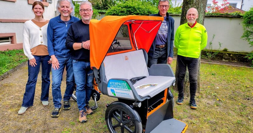 Nicole Novak, Manfred Rachut, Horst Geißler, Bernd Rupp und Alois Ittner stehen draußen neben dem Gebäude der Stiftung Soziale Gemeinschaft Riedstadt. Vor ihnen steht die Fahrrad-Rikscha mit orangem Verdeck und grauen Sitzpolstern sowie grauer Fußbank.